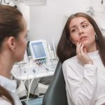 A woman in a dental chair touches her cheek while talking to a dentist in a clinic setting.